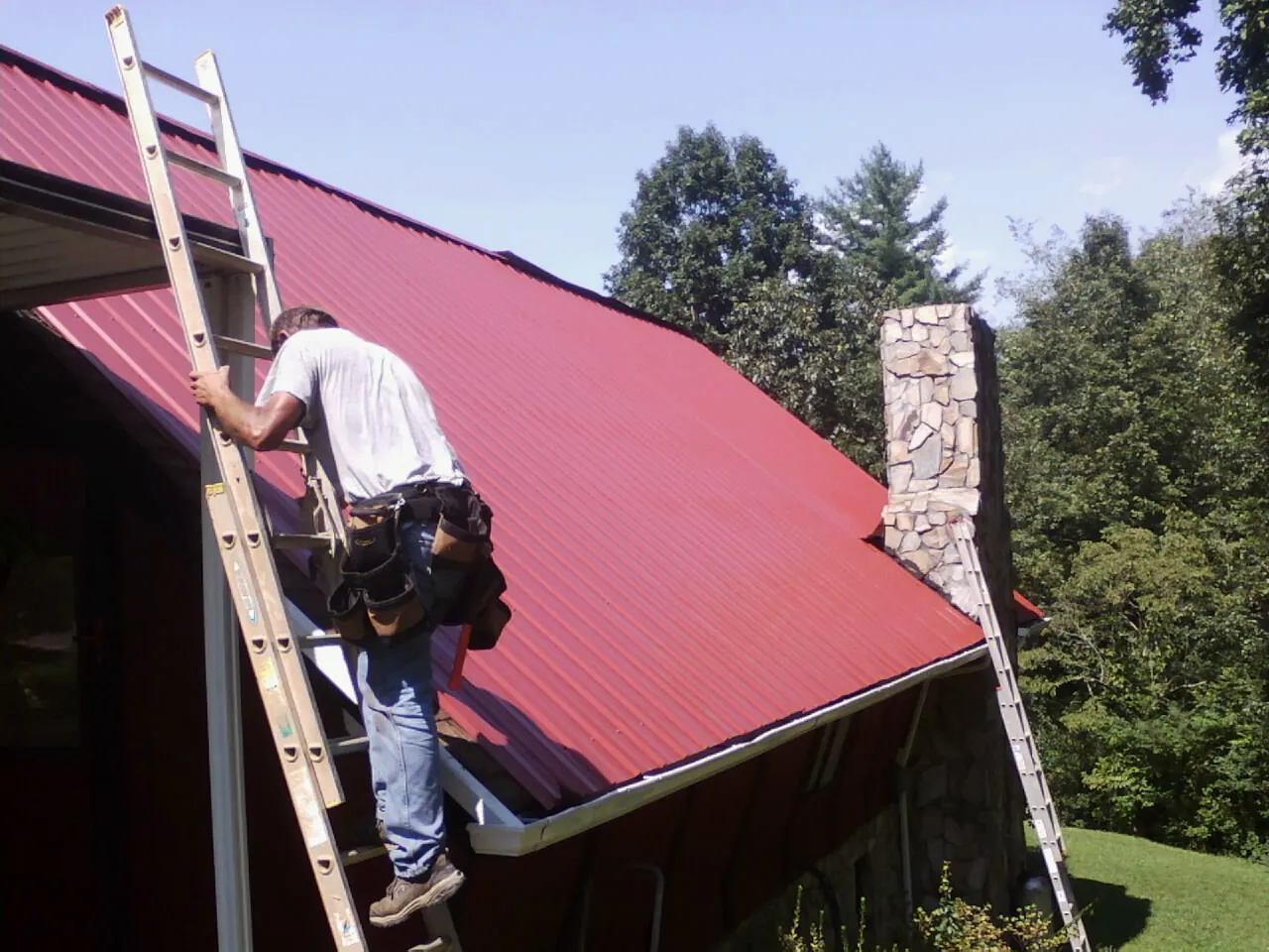 Roofer on a ladder beside a red metal roof and stone chimney — Soffit & Fascia Repair by Roofing Brothers in Fishers Island